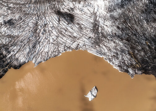 Aerial view of the edge of a glacier with deep crevasses and dark sediment meeting silty brown water in Vatnajokull National Park, Iceland.