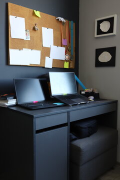 Vertical shot of modern home office interior with two laptops on dark desk. Cozy remote work space, cork board with paper notes pinned on grey wall, corporate freelance workplace architecture
