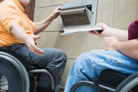 Couple in wheelchairs with Muscular Dystrophy returning a book in a library slot