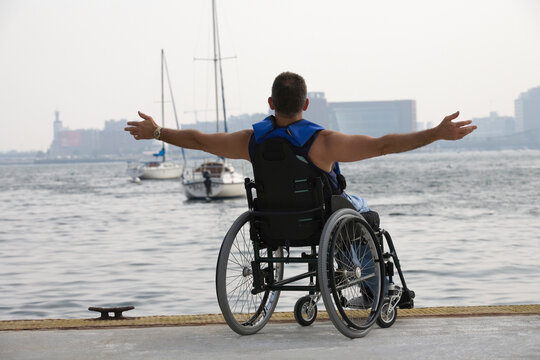 Rear view of a man sitting in a wheelchair with his arm outstretched at a harbor