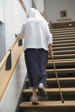 Rear view of a nun moving up on a staircase with the help of a cane