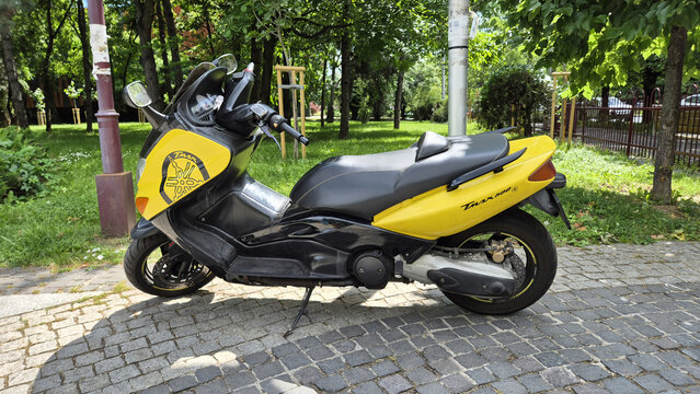 A yellow and black Yamaha TMAX 500 maxi-scooter parked on a cobblestone pathway near a park with green trees. Picture taken on June 8, 2026 in Bucharest, Romania.