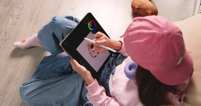 Child wearing pink cap drawing cute cartoon capybara on digital tablet screen with stylus pen. Creative educational graphic design software activity for early child development at home. Real indoor