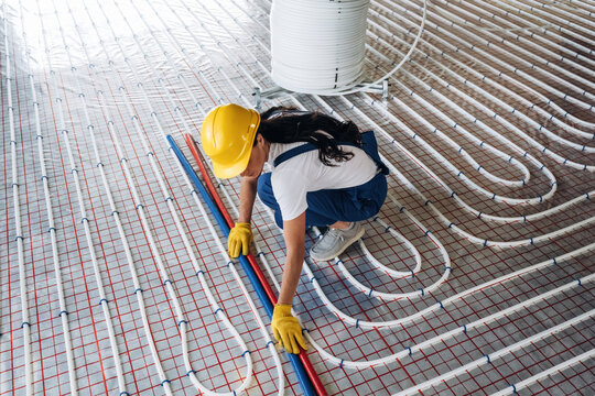Female construction worker in yellow hard hat and gloves installs heating pipes on floor with grid pattern in a modern building under construction