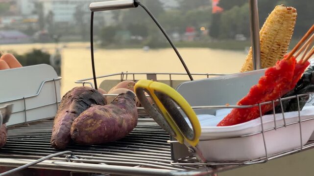 Street vendor roasts sweet potatoes over charcoal on a portable grill near a lakeside promenade with visible urban buildings across the water. Street food.