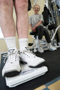 Husband standing on weighing machine with wife sitting in the background