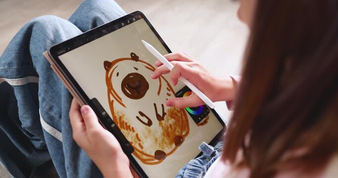 Child wearing pink cap drawing cute cartoon capybara on digital tablet screen with stylus pen. Creative educational graphic design software activity for early child development at home. Real indoor