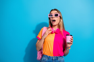 Fototapeta premium Young woman in colorful outfit with pink headphones holding coffee on blue background lifestyle model