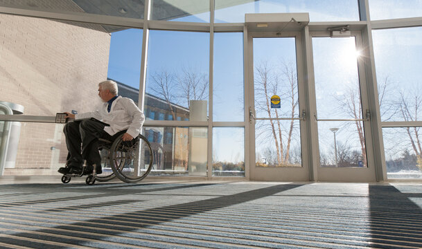 Doctor with muscular dystrophy in wheelchair at hospital entrance pressing accessible door plate