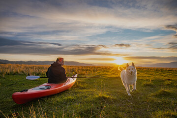Man with paddle sitting in red kayak on green grass while white dog runs during golden hour. Traveler with husky enjoying outdoor adventure in scenic valley under cloudy sky. © ANDREY