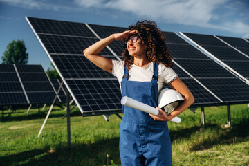 Naklejka premium Female engineer with curly hair wearing blue overalls and glasses stands in front of solar panels, holding a helmet and rolled blueprints, shielding eyes from sunlight