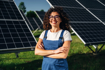 Female technician with curly hair in blue overalls stands confidently with arms crossed in front of solar panels on a sunny day in a green field © standret