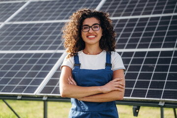 Female technician with curly hair wearing blue overalls stands confidently in front of solar panels on a sunny day, showcasing renewable energy technology in a green field © standret
