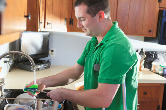Man after anterior cruciate ligament (ACL) surgery with crutches washing dishes in the kitchen