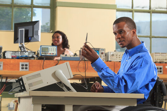 Engineering students in an electronics classroom, one in a wheelchair examining an oscilloscope