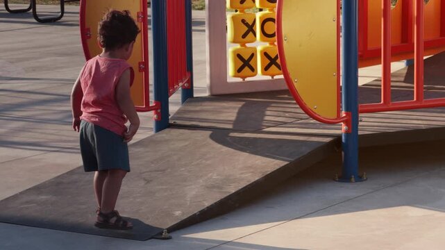 Cute little boy in pink vest and blue shorts runs on slope at outdoor public playground during golden hour - released for commercials
