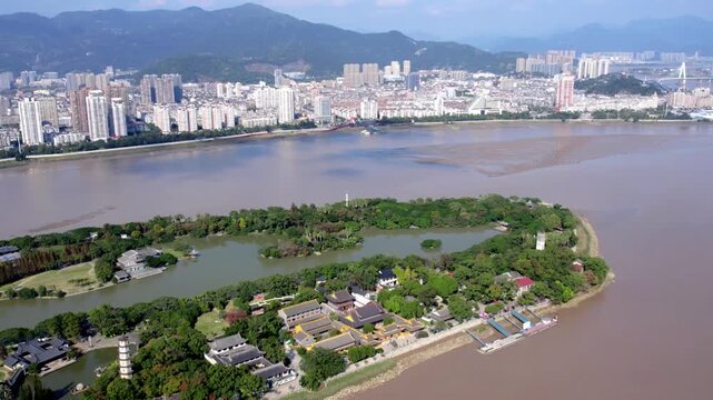 Aerial view of a tree-covered island with ancient architecture, set against a modern urban skyline across a muddy river with distant mountains.