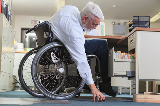 Man with Muscular Dystrophy in a wheelchair reaching for something he dropped at his office desk