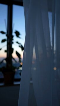 Sheer white curtains gently sway in the evening breeze, revealing a blurred city skyline and potted plants against a twilight backdrop of soft blue and orange hues