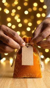 Hands of a person tying a decorative string around a small orange velvet pouch with a blank tag, set against a backdrop of warm, glowing lights