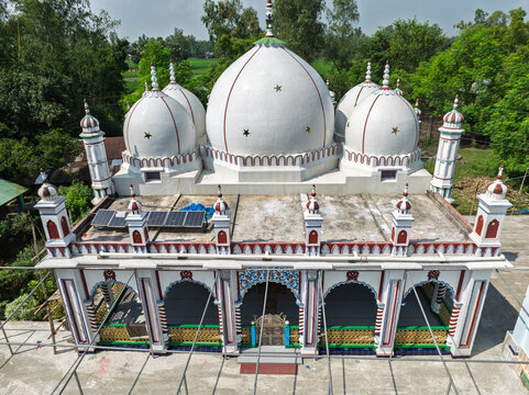 Aerial view of a white mosque with multiple domes and minarets featuring solar panels on the roof and surrounded by lush green trees in Joypurhat, Rajshahi Division, Bangladesh.