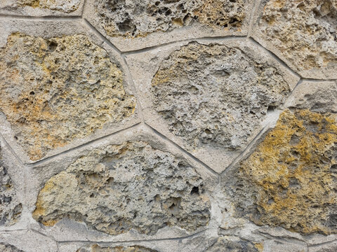 Close-up of a rustic stone wall texture featuring porous, volcanic or coquina stones in shades of grey and yellow, held together by clean concrete mortar joints.