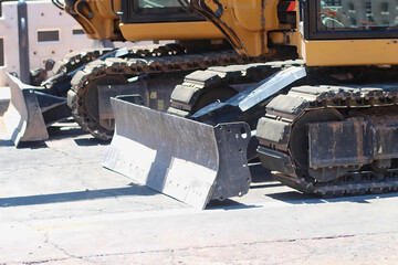 Close-up of tracked construction vehicles lined up on worn pavement, featuring heavy steel blades and rugged treads under bright daylight in an urban work setting. © Priscilla