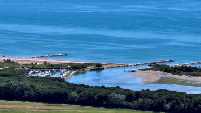 Wide aerial drone tracking shot revealing the small maritime port channel entrance and boat docks near the Piave river mouth and Eraclea lagoon coast in bright daylight, Italy