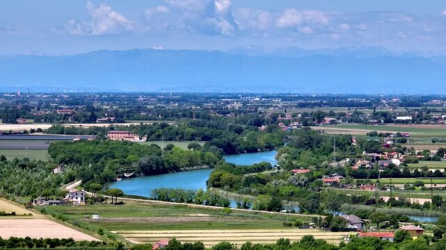 High wide drone panorama following the winding loop of the Piave river flowing through the green Venetian plain towards the distant mountains under a blue sky, Eraclea, Italy