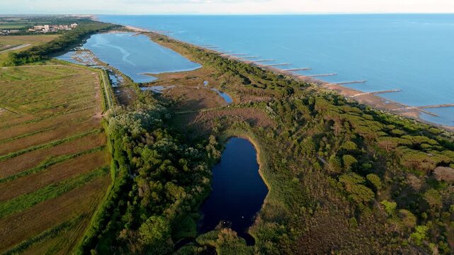 Wide aerial drone perspective of the narrow coastal spit separating the calm sea waves from the wild lagoon marshes and inner channels at sunset near Eraclea and the Piave river, Italy