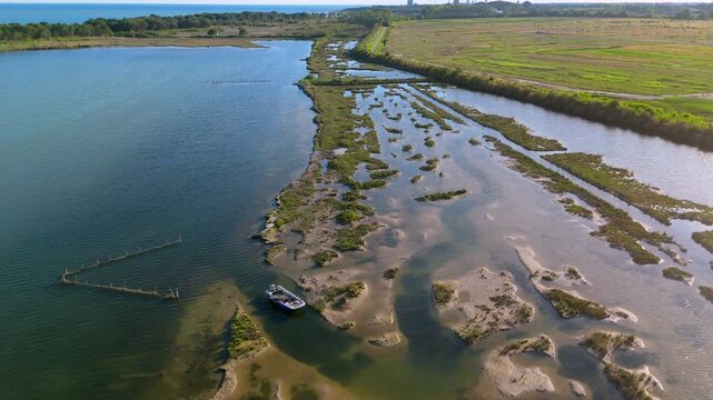 drone overflight following a narrow blue channel winding between wild green marsh vegetation and mudflats in the Venetian Lagoon near Eraclea at sunset, Veneto, Italy