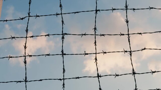 Barbed wire fence creating a grid pattern against a clear blue sky with soft white clouds, symbolizing concepts of security, protection, confinement, borders, and restriction