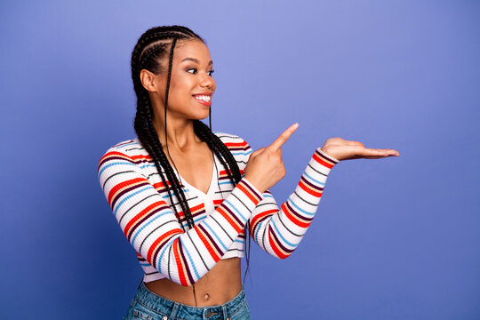 Young stylish woman with braids points to her open hand against a blue background wearing a striped sweater and jeans