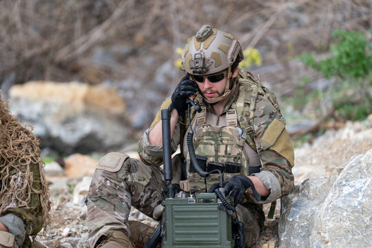 Soldier in camouflage uniform talking on tactical military radio transceiver station outdoor, special forces operator using communication equipment near rock cliff background
