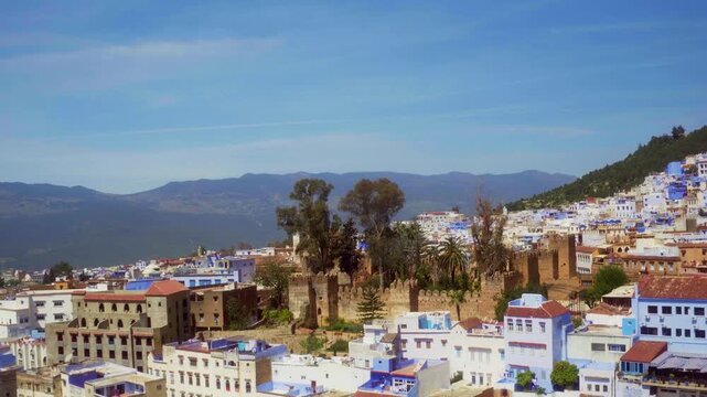 Drone retreats from Chefchaouen kasbah showing blue medina, surrounding city, and Rif valley.