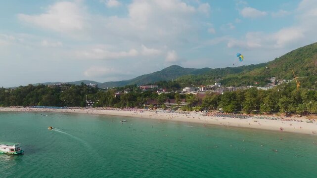 Drone tracks parasail over Patong beach, turquoise water, resort town, and tropical hills.