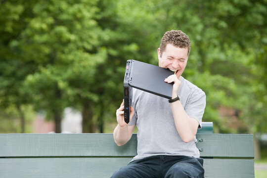 Mid adult man sitting on a park bench and biting a laptop