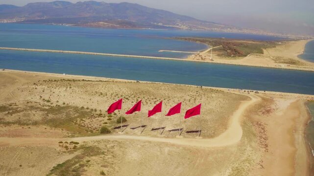 Drone approaches five red flags on arid sandy spit between coastal lagoon and open sea.