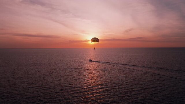 Drone approaches parasailer silhouette towed by boat against vivid orange sunset over calm sea.