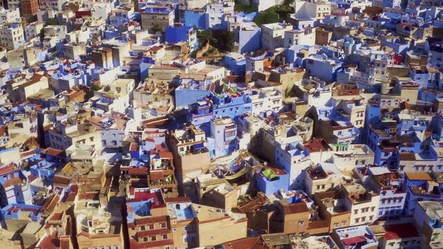Drone pans in steep plunge over Chefchaouen medina, casting deep shadows across blue and white rooftops.