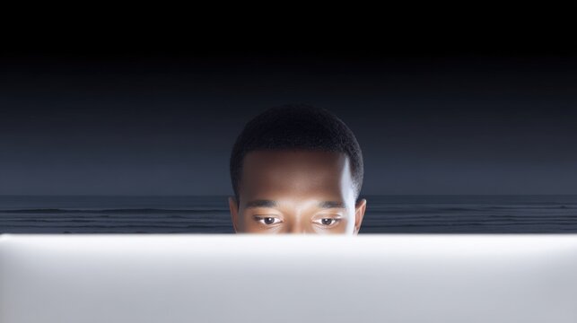 Young person with short hair looking at a white laptop screen with a dark night sky, ocean waves, and beach in the background