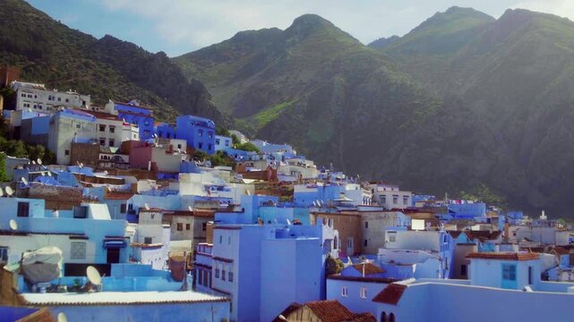 Drone rises from Chefchaouen street over blue rooftops, sun behind Rif peak.