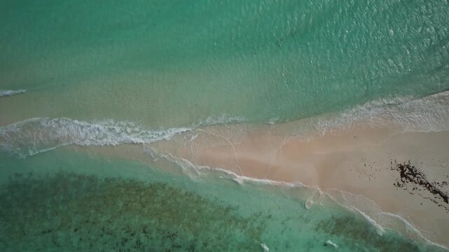 mesmerizing top-down drone capture of gentle turquoise waves washing over the pristine, narrow white sand isthmus of a Caribbean paradise.