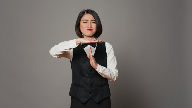 Front desk employee asking for a break in studio, showing timeout symbol and feeling overworked, tired Asian receptionist dealing with burnout and asking for a pause, against a grey studio background