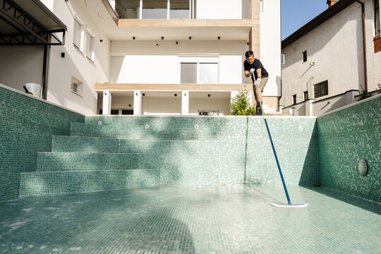 Cleaning an Empty Swimming Pool Before Seasonal Startup With a Technician Using a Pole and Scrubbing Tools