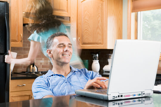 Mid adult man sitting in a wheelchair and using a laptop with a mid adult woman walking behind him