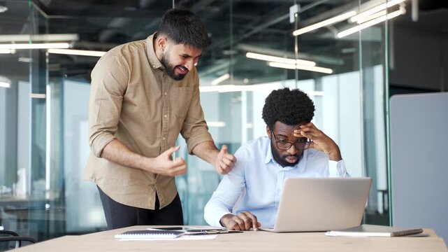 Furious boss standing in modern office yelling at employee sitting at desk with laptop. Male manager blaming subordinate, expressing anger and stress during conflict, arguing about work mistake