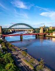 Fototapeta premium A vibrant shot of a steel arch bridge and a red bridge spanning a wide river under a bright blue, cloud-dotted sky