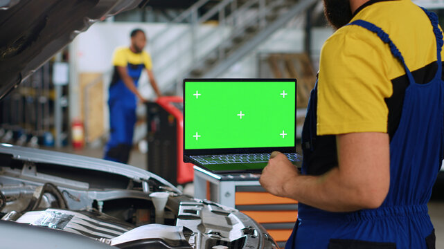 Licensed repairman using green screen laptop in car service to order new parts for busted vehicle. Close up shot of professional using chroma key device to look on internet for replacing components.