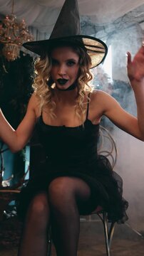 Beautiful young sorceress in a black witch hat and dress, smiling alluringly while casting a glowing spell amid smoky, mysterious halloween party ambiance. Vertical video